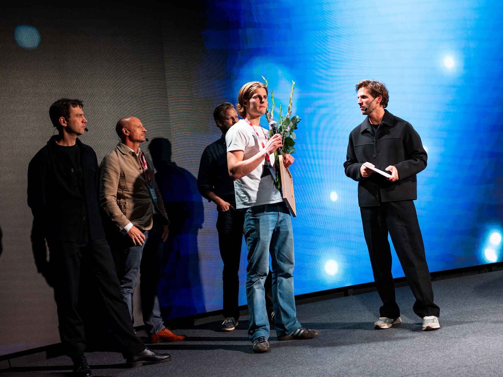Gustav on the AI x SaaS Summit stage with flowers and the diploma, next to the host and the other winners